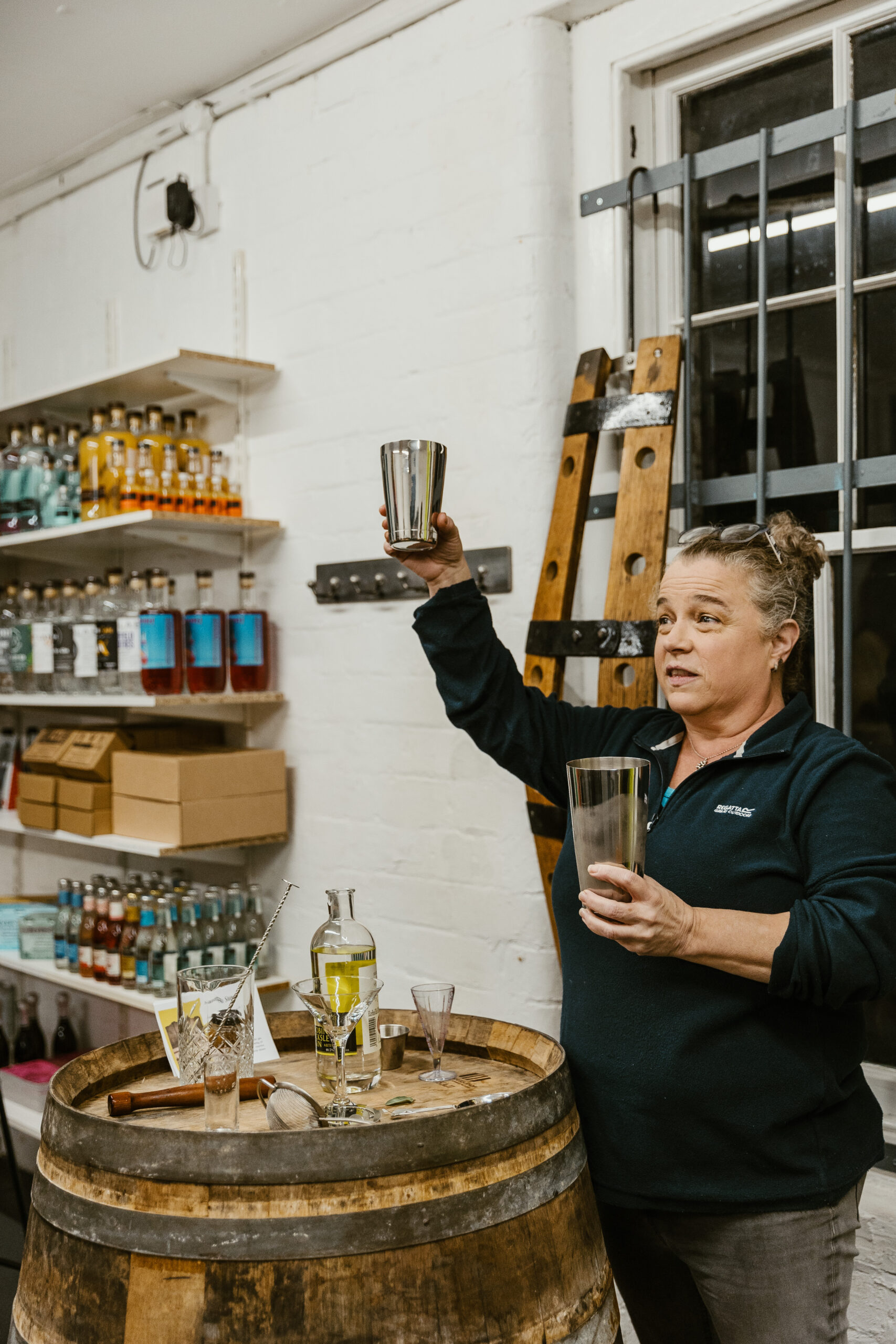 A short white women with greyish hair pulled back, holding one part of a cocktail shaker in each hand, one high in the air